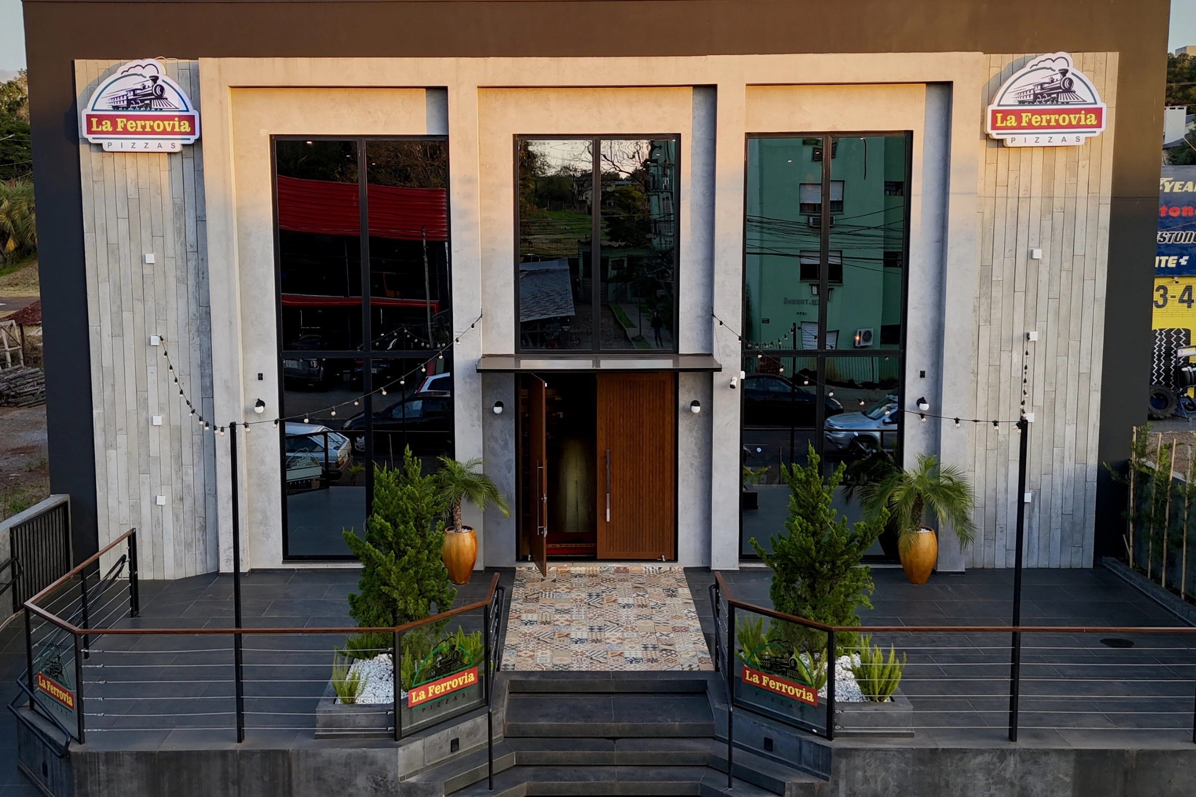 Fachada de um edifício comercial moderno com grandes janelas de vidro e detalhes de madeira na porta de entrada, ladeado por plantas e com iluminação externa.